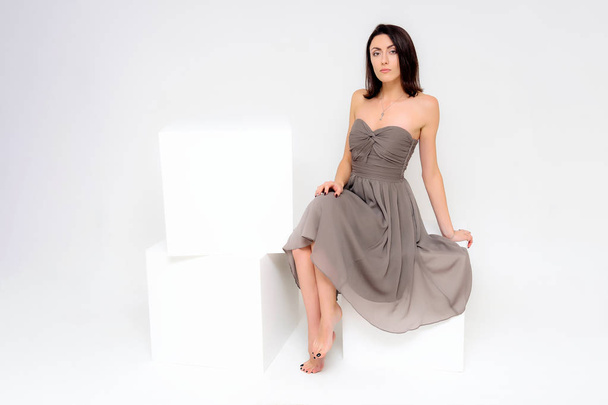 Concept portrait of a brunette girl, sitting on a white background in a beautiful brown dress on a white cube in studio. She sits right in front of the camera in various poses with different emotions. - Fotoğraf, Görsel