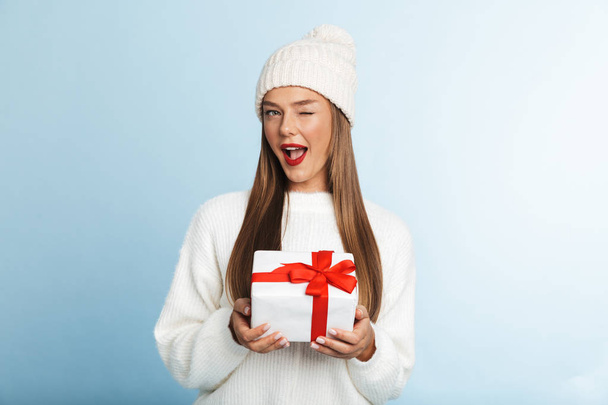 Cheerful young woman wearing sweater and hat isolated over blue background, holding gift box, winking - Valokuva, kuva