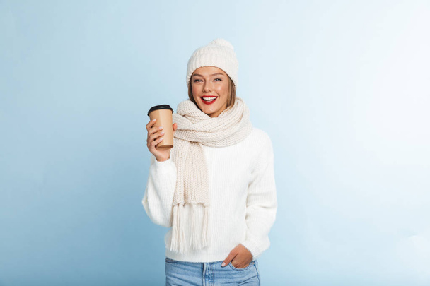 Happy young woman wearing sweater and hat isolated over blue background, holding takeaway coffee - Photo, Image