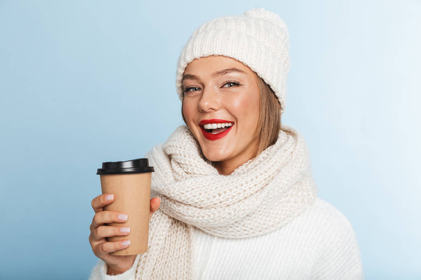Happy young woman wearing sweater and hat isolated over blue background, holding takeaway coffee - Fotografie, Obrázek