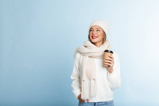 Happy young woman wearing sweater and hat isolated over blue background, holding takeaway coffee - Фото, изображение