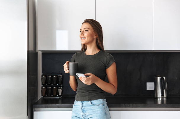 Happy young woman standing at the kitchen, drinking tea, using mobile phone - Zdjęcie, obraz