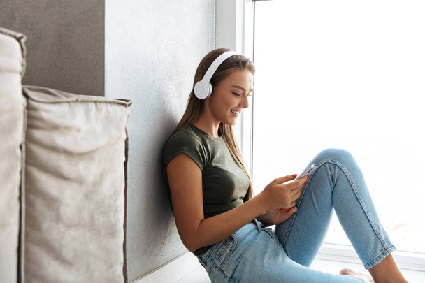 Cheerful young woman listening to music with headphones at home, using mobile phone - Photo, Image