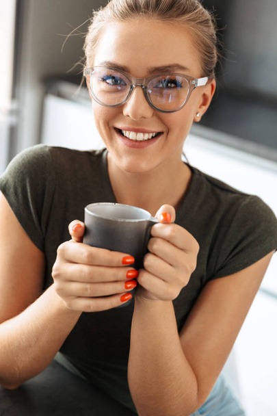 Smiling young woman holding a cup while sitting at the kitchen - Fotografie, Obrázek
