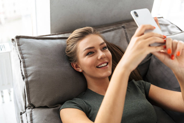 Cheerful young woman taking a selfie while laying on a couch at home - 写真・画像
