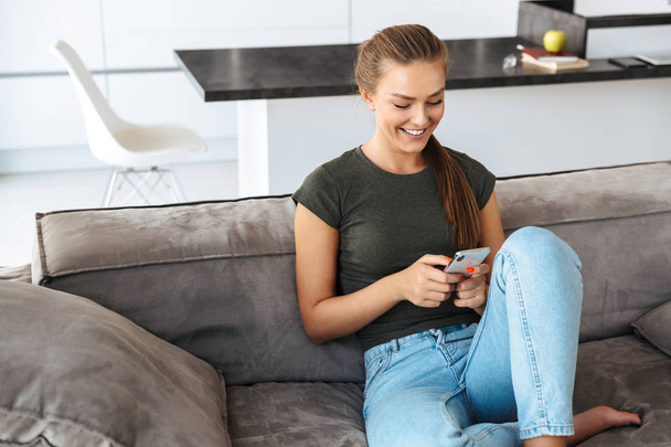 Cheerful young woman using mobile phone while sitting on a couch at home - 写真・画像