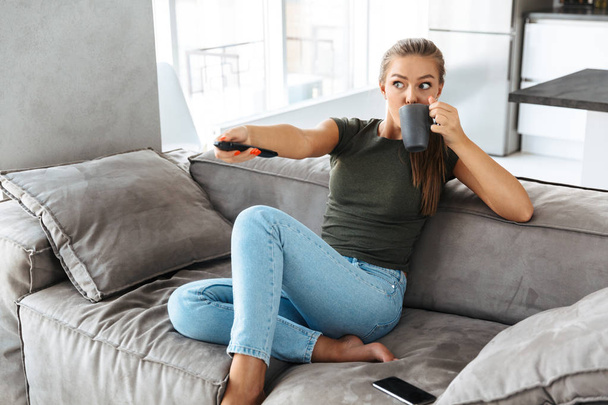Cheerful young woman sitting on a couch at home, changing channels, drinking tea from a cup - Valokuva, kuva