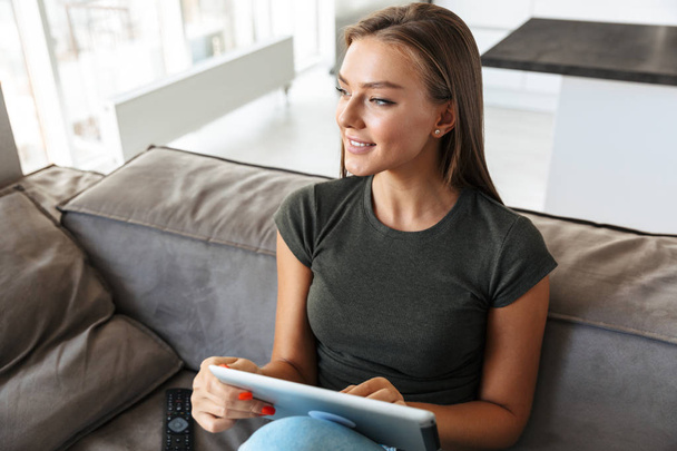 Smiling young woman sitting on a couch at home, using tablet computer - Fotó, kép