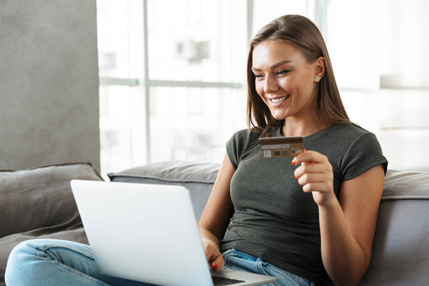 Smiling young woman sitting on a couch at home, using laptop computer, showing credit card - Fotografie, Obrázek