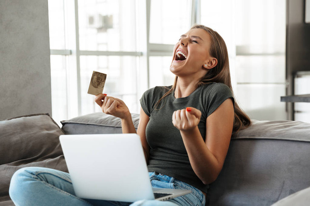 Smiling young woman sitting on a couch at home, using laptop computer, showing credit card - Fotoğraf, Görsel