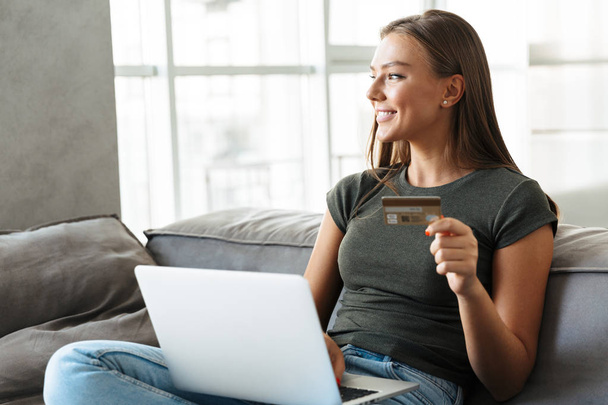 Smiling young woman sitting on a couch at home, using laptop computer, showing credit card - Foto, Imagem