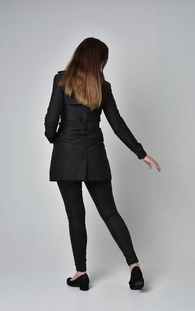 täyspitkä muotokuva brunette tyttö yllään pitkä musta takki, seisoo poseeraa takaisin kameran harmaa studio tausta
. - Valokuva, kuva