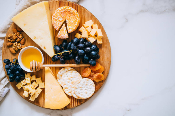 Acostado plano con varios tipos de queso, uvas, nueces, miel y galletas en tablero de madera sobre mármol
 - Foto, Imagen