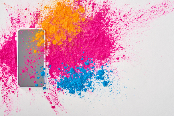 vista superior del teléfono inteligente y explosión de polvo de holi multicolor sobre fondo blanco
 - Foto, Imagen