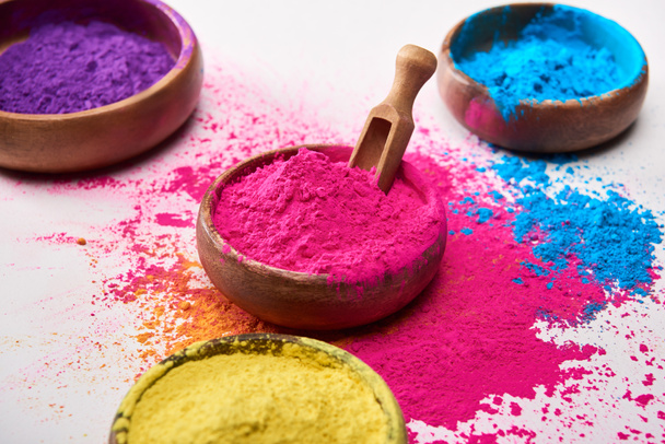 wooden spatula and bowls with yellow, pink, blue and purple holi powder on white background - Photo, image
