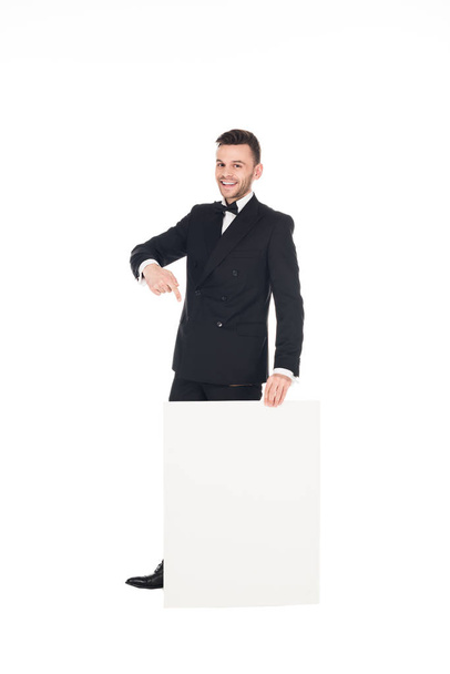 cheerful elegant man in black suit pointing at empty board isolated on white - Foto, Imagem