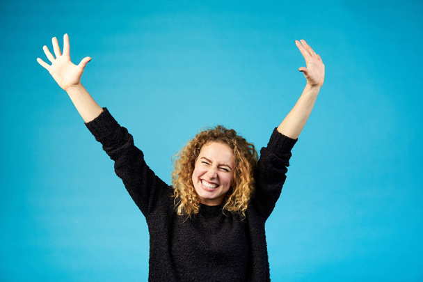 Happy satisfied young ginger curly woman celebrating and cheering a success spreading hands with joy against blue background. Positive feelings. Studio shot. - Photo, Image