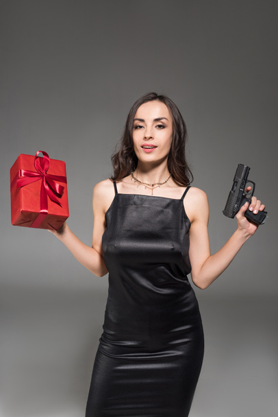 mooie brunette vrouw in zwarte jurk rode luxe-geschenketui en pistool geïsoleerd op grijs - Foto, afbeelding