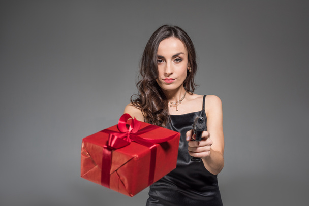 serious brunette woman holding red present and gun isolated on grey - Photo, Image