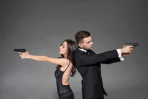 confident couple of secret agents in black clothes aiming with weapon, isolated on grey - Foto, imagen