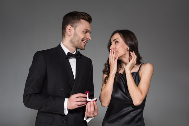 handsome boyfriend holding proposal ring in box for excited girlfriend, isolated on grey - Fotografie, Obrázek