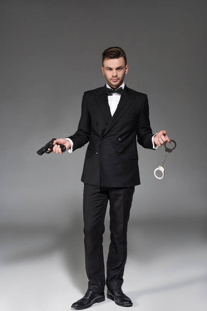 elegant secret agent in tuxedo holding handgun and handcuffs on grey - Foto, Imagen