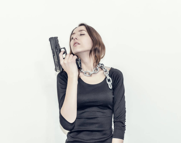 Slim figure girl in the black dress. Metal chain around the neck. Black gun in hand. Slavery, exploitation of people, lack of freedom, violence, threat, liberation. - 写真・画像