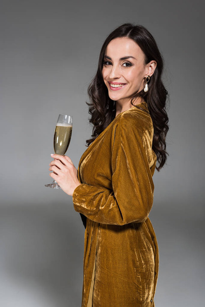 beautiful smiling woman in golden dress holding glass of champagne isolated on grey - Zdjęcie, obraz