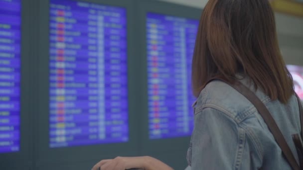 Cámara lenta - Mujer asiática feliz mirando el tablero de información que comprueba su vuelo con el equipaje en el pasillo de la terminal en la puerta de salida en aeropuerto internacional. Estilo de vida de las mujeres felices en concepto de aeropuerto
. - Imágenes, Vídeo