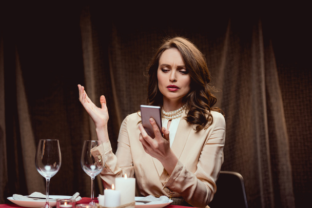 beautiful confused woman sitting at table and gesturing while using smartphone in restaurant - Photo, Image