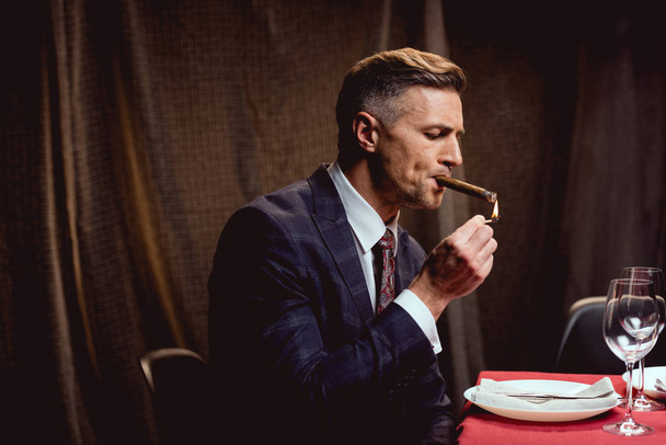handsome man in suit sitting at table and lighting up cigar in restaurant - Zdjęcie, obraz