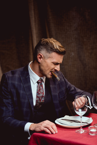 insatisfait bel homme en costume assis à la table, fumer le cigare et regarder regarder dans le restaurant
 - Photo, image