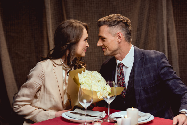 hermosa pareja sentada en la mesa con ramo de flores y mirándose durante una cita romántica en el restaurante
 - Foto, imagen