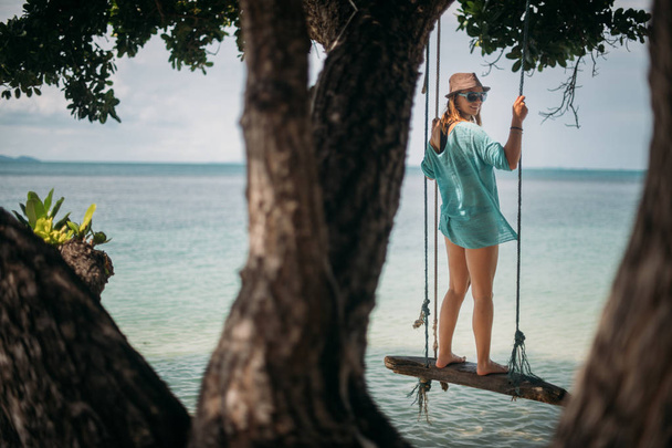 Een jonge vrouw is swingend op een schommel in de schaduw van een grote boom door de zee. Meisje op de schommel strand aan de kust van een tropisch eiland. - Foto, afbeelding