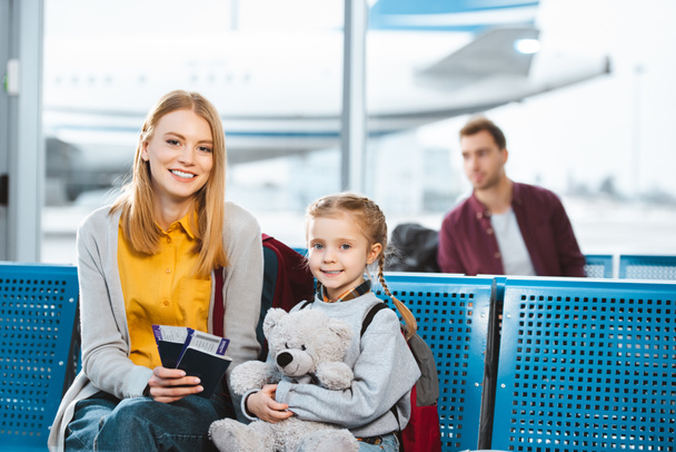 selective focus of mother holding passports with air tickets and smiling near daughter in airport in departure lounge - Photo, Image