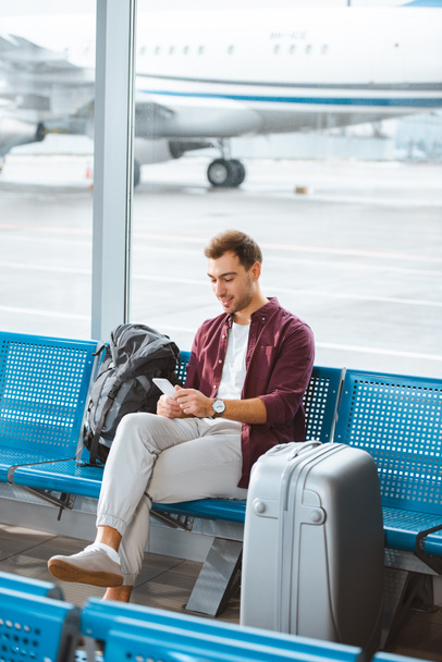 smiling man using smartphone while sitting in waiting hall in airport - Photo, Image