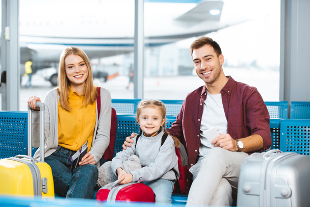 happy family sitting in departure lounge and smiling near luggage in airport - Foto, Imagem