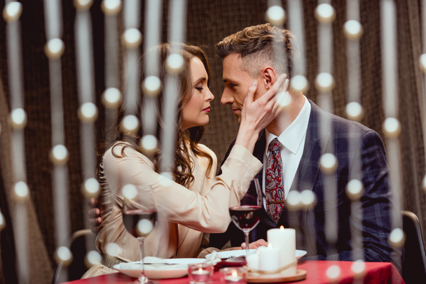 Kobieta, delikatnie dotykając twarz człowieka podczas romantyczną randkę w restauracji z bokeh świateł na pierwszym planie - Zdjęcie, obraz