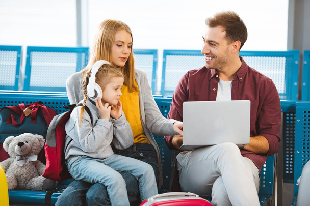 приваблива жінка розмовляє з чоловіком з ноутбуком біля дочки в навушниках в аеропорту
  - Фото, зображення