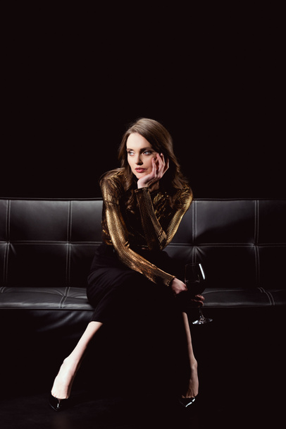 hermosa mujer glamorosa sentada en el sofá con copa de vino tinto y barbilla de apoyo aislado en negro
 - Foto, imagen
