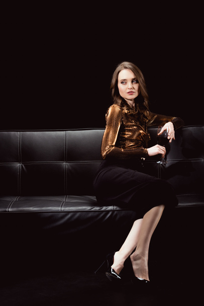 hermosa mujer glamorosa sentada en el sofá con una copa de vino tinto y mirando hacia otro lado aislado en negro
 - Foto, Imagen