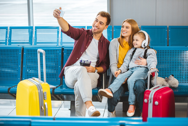 família alegre levando selfie e sorrindo no aeroporto perto de bagagem
 - Foto, Imagem