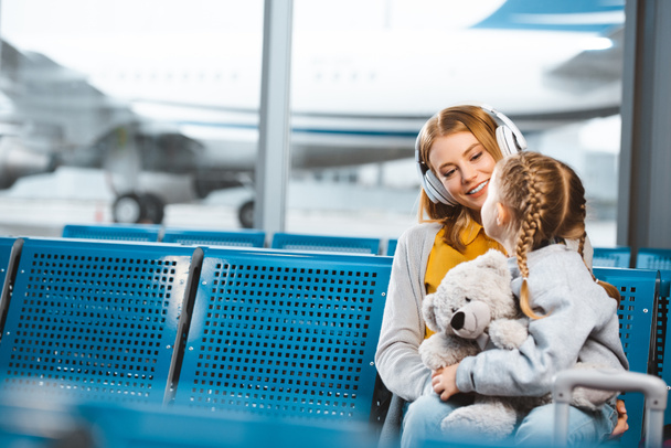 beautiful mother in headphones looking at daughter with teddy bear in departure lounge - Photo, Image