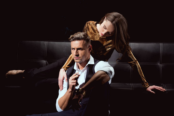 schöne Frau liegt auf dem Sofa und umarmt gutaussehenden Mann mit Zigarette isoliert auf schwarz - Foto, Bild