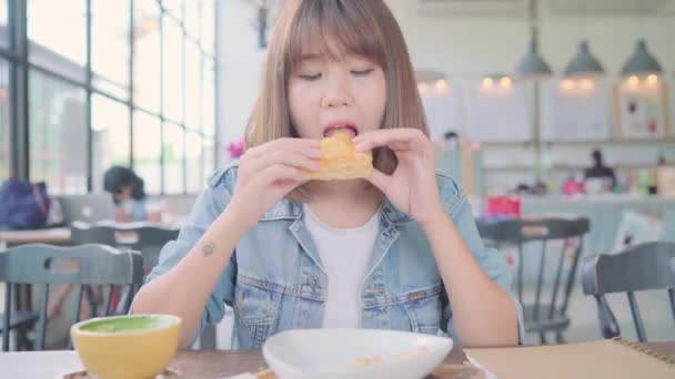 Freelance Aziatische zakenvrouw eten brood en het drinken van warme kop koffie zittend aan tafel in café. Levensstijl slimme mooie vrouw ontspannen in koffie shop concepten. - Video