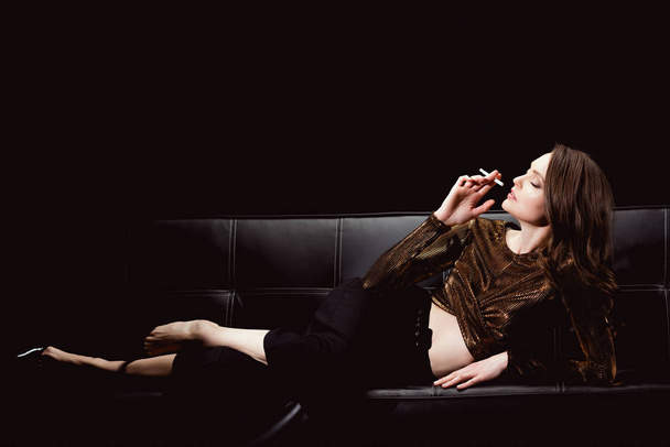 beautiful glamorous woman lying on couch and smoking cigarette isolated on black - Photo, Image