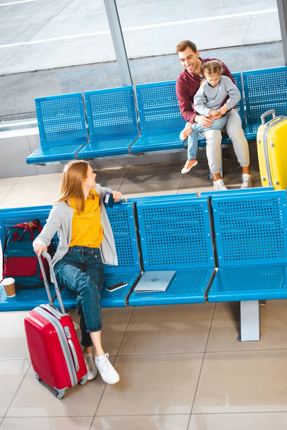 ευτυχής πατέρας κάθεται με κόρη στο αεροδρόμιο κοντά γυναίκα με αποσκευές  - Φωτογραφία, εικόνα