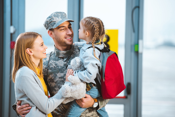 smiling father in military uniform holding in arms daughter near wife in airport  - 写真・画像