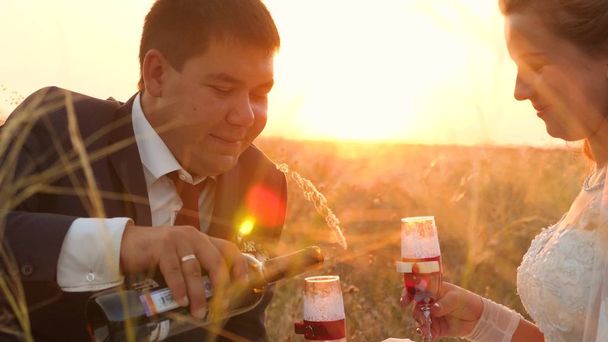 Bräutigam schenkt köstliche Weingläser in Feld in hellen Sonnenstrahlen für Braut. Nahaufnahme. Das glückliche Brautpaar trinkt Champagner auf der Wiese im Sonnenuntergang. Konzept der glücklichen Hochzeit - Foto, Bild