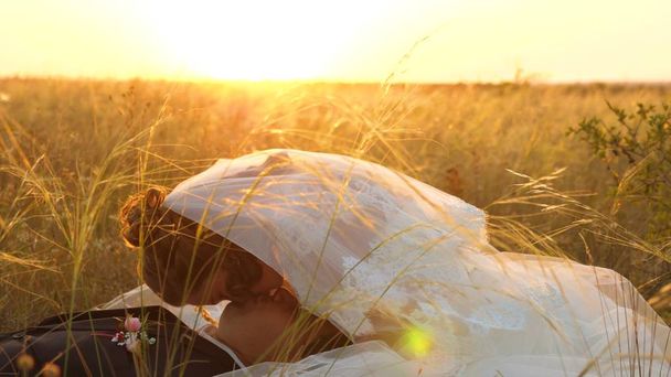 bride caresses groom with grass stalk in face. happy family is lying in field in rays of beautiful sunset. young woman kisses her beloved man on lips on summer evening in the grass. happy family - Photo, Image